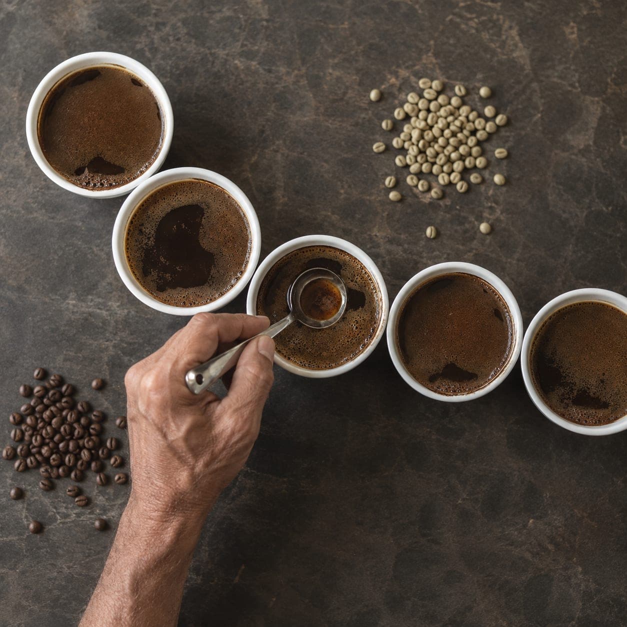 Overhead view of a specialty cupping table with porcelain bowls of brewed coffee, a working hand holding a silver cupping spoon, and piles of beans.